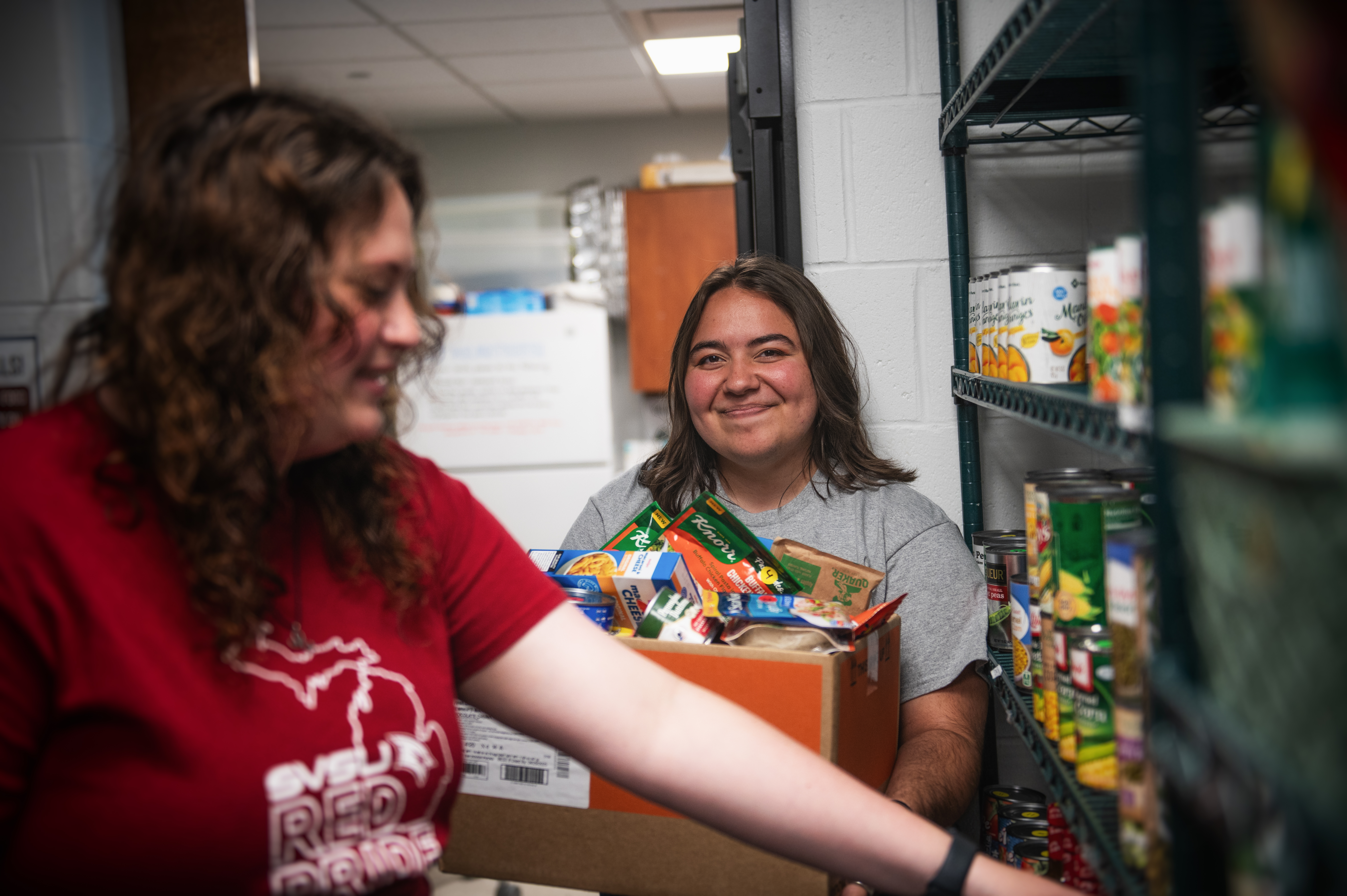 Student looking at the camera while holding a box of groceries in the food pantry while another student is shopping and taking items from the shelves.